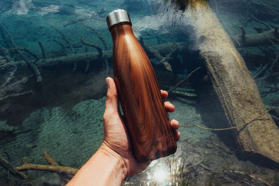 Une main tient une gourde filtrante au motif boisé devant une eau de lac transparente.