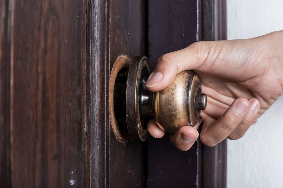 Une main actionne une poignée de porte ronde en laiton fixée sur une porte en bois sombre, représentant un cas de carré de poignée de porte cassé ou usé.